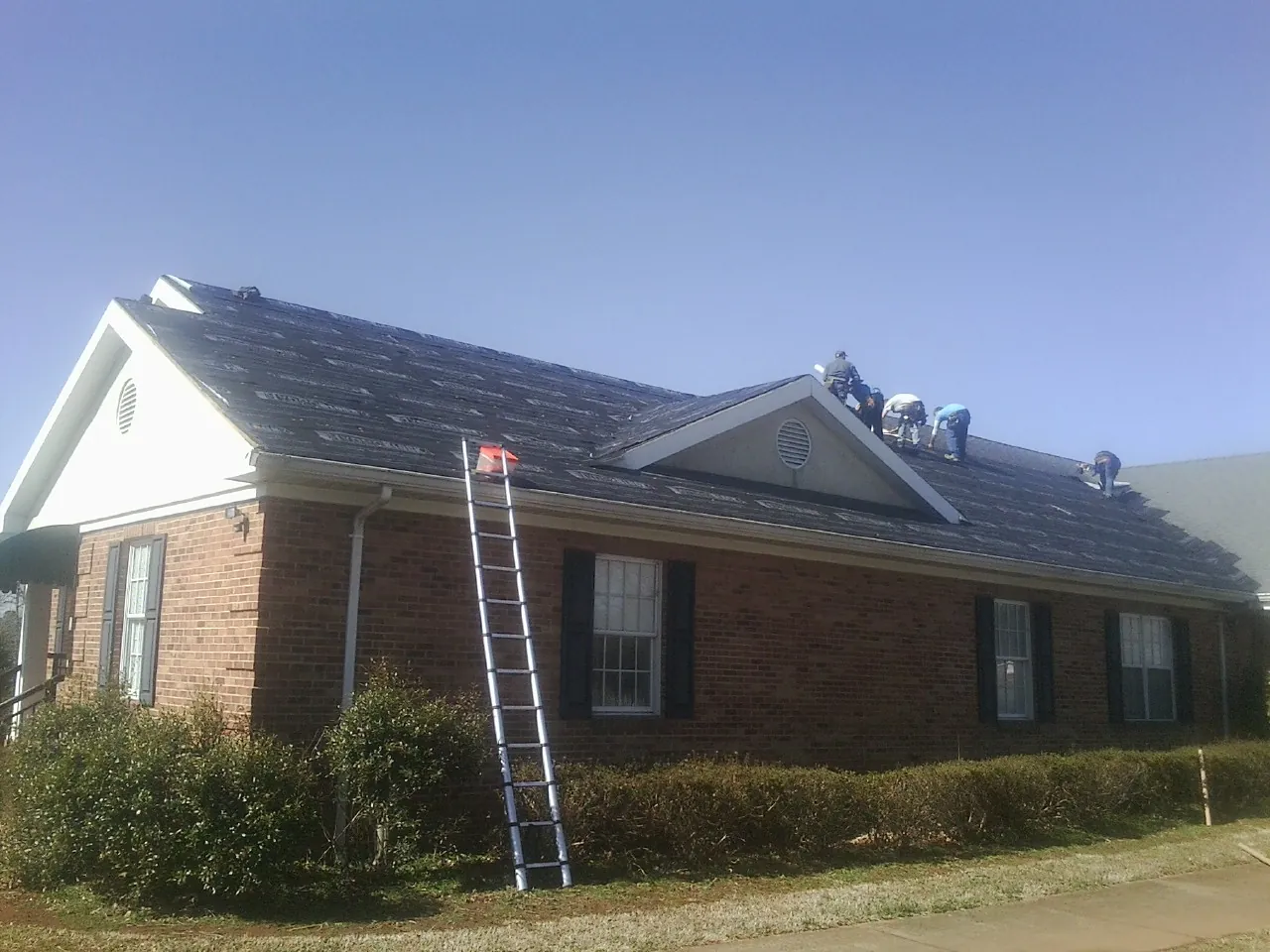 Crew tearing shingles off a brick ranch roof with a ladder set — Emergency Roof Repair work by our crew in Mount Dora
