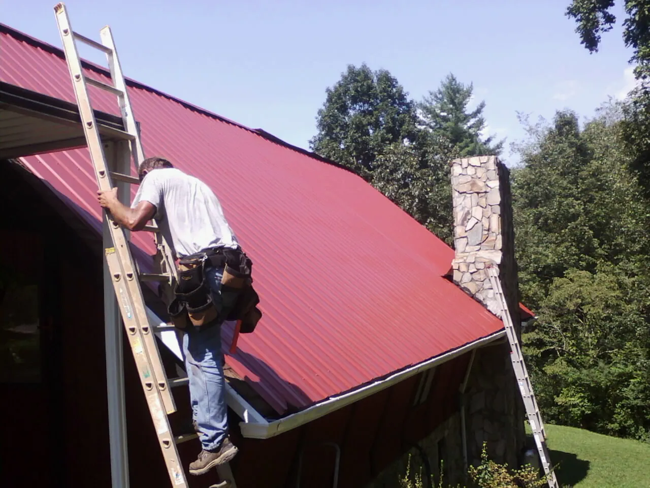 Roofer on a ladder beside a red metal roof and stone chimney — Soffit & Fascia Repair by Roofing Brothers in Mount Dora